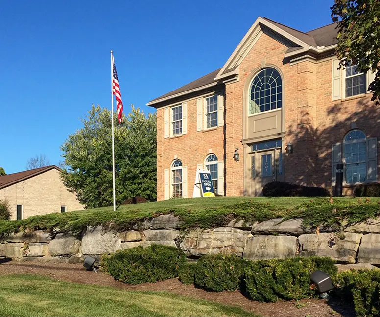 Brick S&A Homes sales office with arched windows, stone retaining wall, landscaped grounds, and American flag.